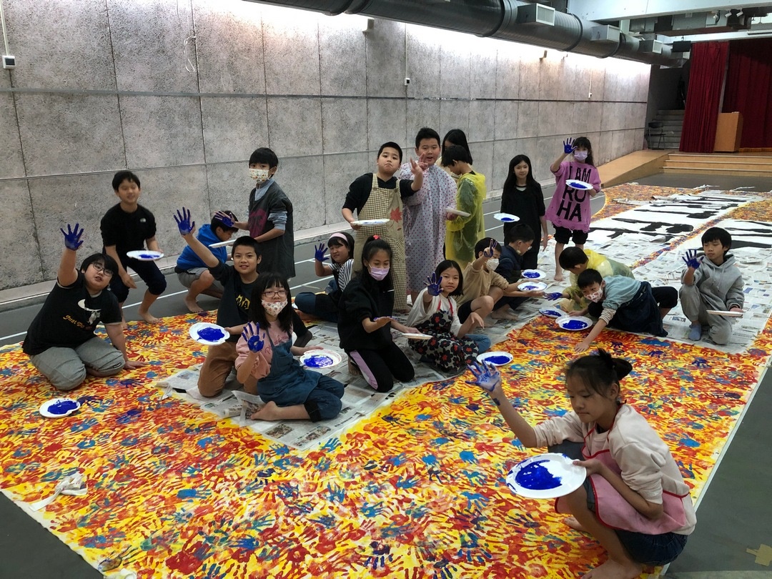 學生們席地而坐，手掌沾滿藍色顏料，開心地揮舞著雙手，準備在巨幅畫布上蓋下屬於自己的印記。鼓勵學生勇敢表達、共同參與，並以藝術為橋樑，凝聚班級與校園的團隊精神，是一場充滿創意與感動的